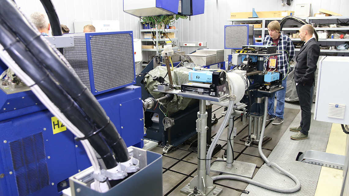 Industrial laboratory test bench with blue machinery, connected instruments, and two people observing the setup during a technical inspection.