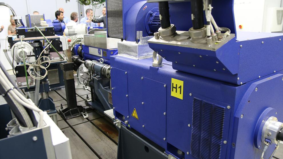 Industrial laboratory setup with large blue experimental machinery and connected mechanical components inside a research facility, with people in the background during a technical demonstration.